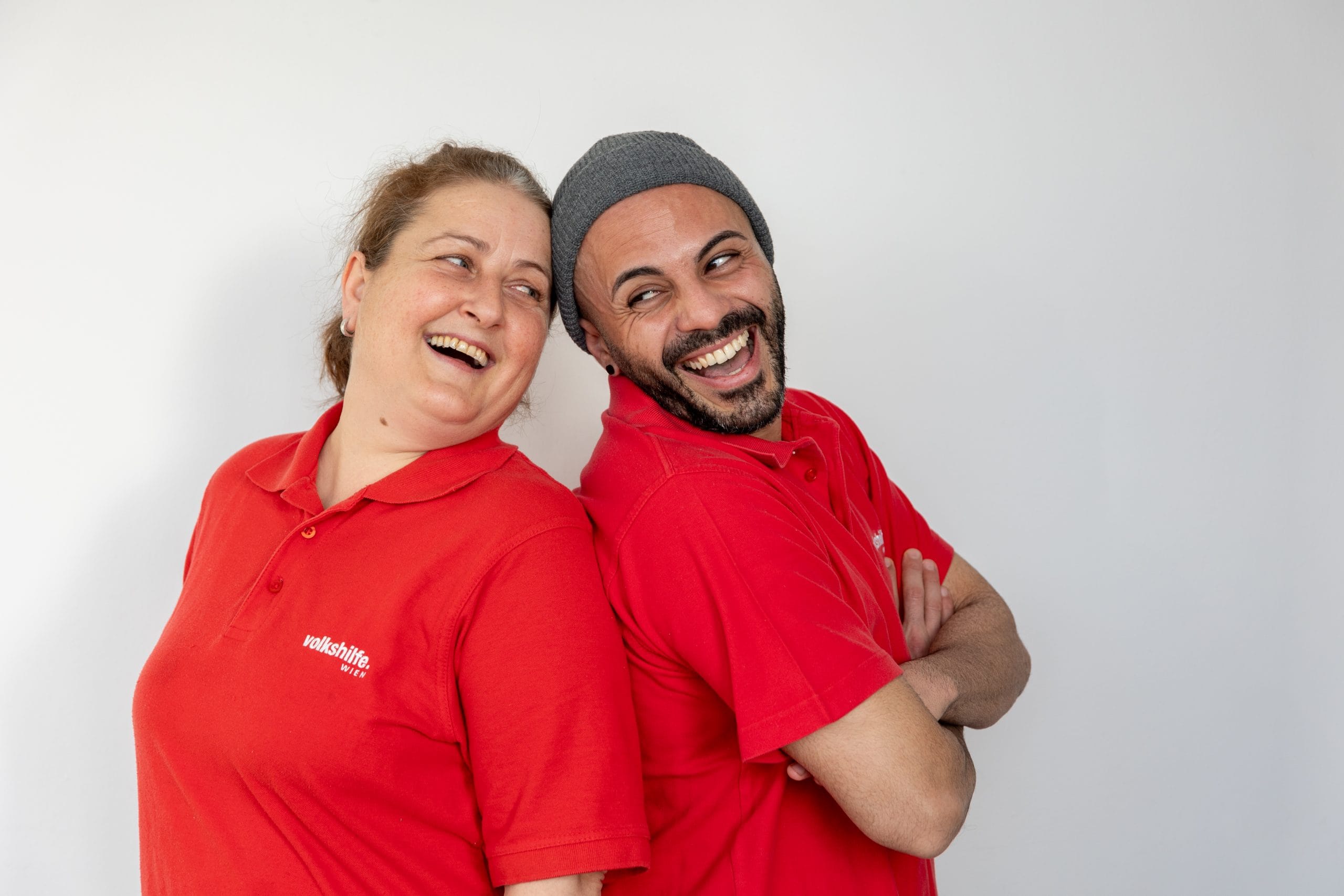 DGKP Zwei DGKPs stehen Rücken an Rücken und lächeln. Beide tragen ein rotes Poloshirt mit der Aufschrift Volkshilfe Wien. Der Hintergrund ist schlicht und weiß.