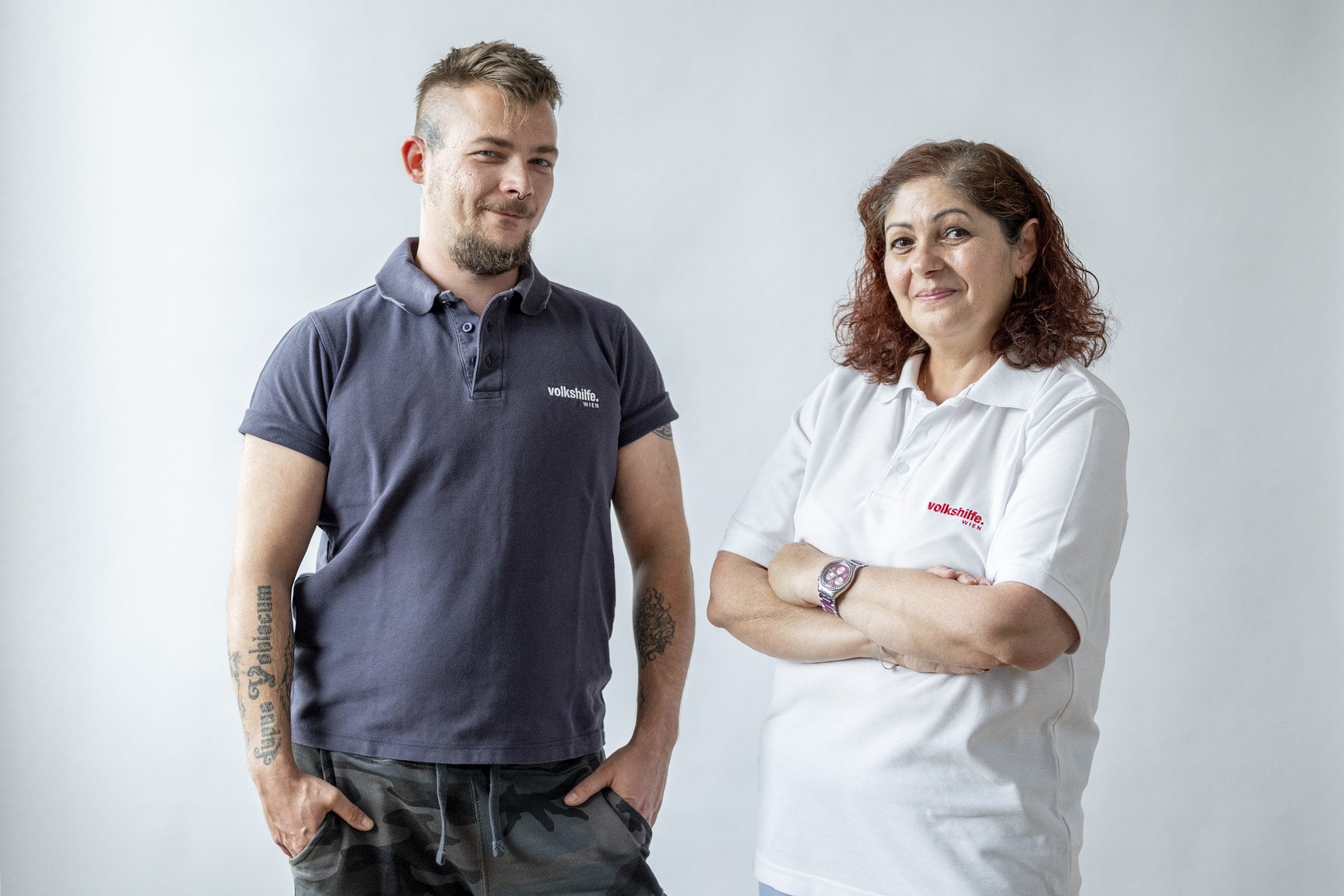 Pflegeassistenz Ein Pflegeassistent steht links und trägt ein dunkelblaues Poloshirt. Eine Pflegeassistentin steht rechts und trägt ein weißes Poloshirt. Auf beiden Poloshirts ist die Aufschrift "Volkshilfe Wien". Beide stehen vor einem schlichten Hintergrund und lächeln.